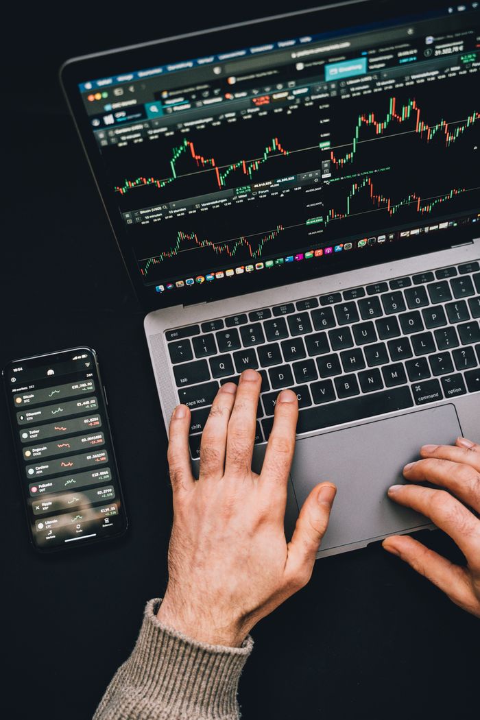 Des mains tapant sur un ordinateur portable avec des graphiques de cryptomonnaies et l’écran d’un smartphone visibles, avec un focus sur la finance numérique.